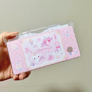 May include: A pink and clear Nintendo Switch case featuring a decorative design with cartoon characters, roses, and the text "Sugarmimuel". The case has a clear top and a pink patterned bottom.