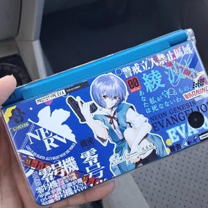 May include: A blue handheld gaming console featuring a graphic of an anime character with blue hair, holding a weapon. The console has Japanese text and the words "NEON GENESIS EVANGELION" and "EVA" printed on it. The console is held in a person's hand.