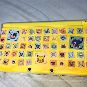 Puede incluir: Una consola de juegos portátil amarilla con una funda protectora transparente decorada con numerosas pegatinas cuadradas pequeñas de varios personajes de dibujos animados coloridos, incluido un icono de Pikachu y un gráfico azul.