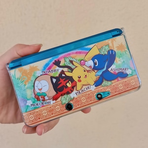 Peut inclure: Une console de jeu portable avec un design coloré mettant en vedette des personnages de dessins animés. La console a un dessus bleu et une coque de protection transparente. Le design comprend les noms "Nyabby", "Pikachu", "Ashimari" et "Mokuroh".