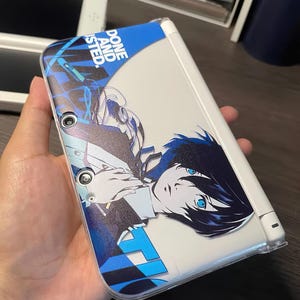 May include: A white handheld gaming console featuring a protective skin with a blue and black graphic design. The skin displays an anime-style character with dark hair and blue eyes, alongside the text "DONE AND ISTED." in a bold white font.