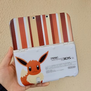 May include: A New Nintendo 3DS LL handheld gaming console featuring a white base with an Eevee character graphic and text, paired with a top cover displaying vertical stripes in shades of brown, cream, and orange, including two camera lenses.