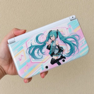 Puede incluir: Una consola de juegos portátil blanca con una cubierta protectora transparente que muestra un personaje de anime con largo cabello color verde azulado, vistiendo un atuendo gris y verde azulado, rodeado de notas musicales y formas.