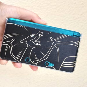 May include: Black handheld gaming console with a teal top and a clear protective case. The console features a white outline graphic of a dragon-like creature. The console is held in a hand.