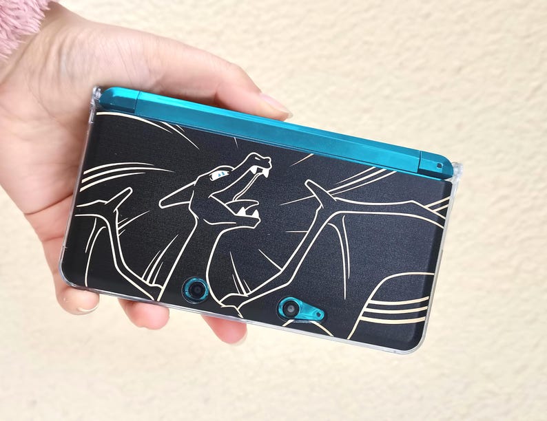 May include: Black handheld gaming console with a teal top and a clear protective case. The console features a white outline graphic of a dragon-like creature. The console is held in a hand.