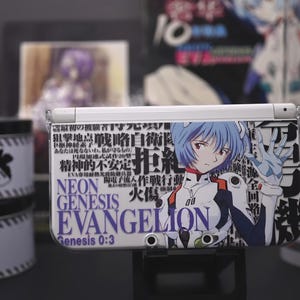 May include: A white Nintendo 3DS handheld game console with a custom cover featuring anime artwork and Japanese text. The cover displays a character with blue hair and the words "NEON GENESIS EVANGELION". Other merchandise is visible in the background.