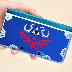以下が含まれることがあります： 赤と白のエンブレムが付いた青い携帯ゲーム機。コンソールは青い上部とシルバーのアクセントが付いています。デザインは人気のビデオゲームシリーズを彷彿とさせます。
