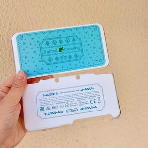 May include: A white Nintendo 3DS console with a light blue cover featuring the "Animal Crossing" logo and a pattern of trees and triangles. The console is open, revealing the interior.