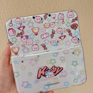 May include: A white handheld gaming console featuring a clear protective case decorated with a repeating pattern of pink Kirby characters, colorful stars, umbrellas, and candies. The back displays the Kirby logo and a small sleeping character graphic.