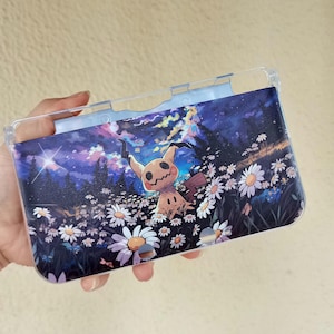 Peut inclure: Une coque de protection en plastique transparent pour console de jeu portable avec une illustration imprimée d'un personnage Mimikyu dans un champ de marguerites blanches sous un ciel nocturne violet et bleu avec une étoile et une forêt.