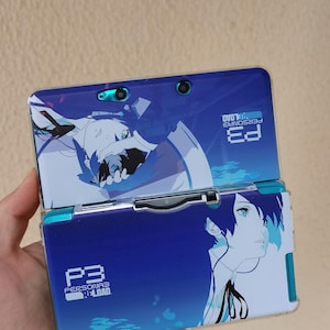 May include: A blue handheld gaming console cover featuring Persona 3 Reload graphics. The top panel displays an anime character with blue hair, while the bottom panel shows a character profile with the text P3 Persona 3 Reload in white lettering.