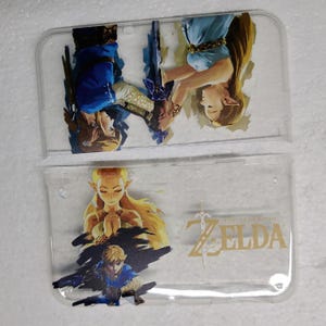 May include: Two clear plastic cases featuring artwork from The Legend of Zelda. One case shows Link and Zelda in a dynamic pose, while the other displays Zelda with the game's logo. The artwork is in shades of blue, gold, and brown.