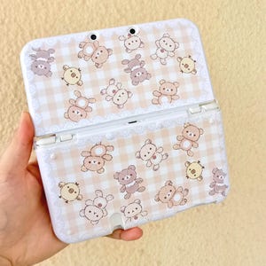 May include: A white Nintendo 3DS case featuring a repeating pattern of cartoon bears and chicks on a light pink and white checkered background. The case has a decorative lace-like border.