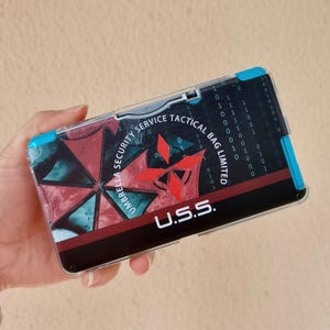 May include: A rectangular protective case for a handheld gaming console featuring a black, red, and teal graphic design. The case displays the text UMBRELLA SECURITY SERVICE TACTICAL BAG LIMITED and U.S.S. with a digital binary code pattern background.