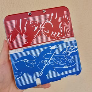 Könnte beinhalten: Eine rote und blaue tragbare Spielkonsole mit einer weißen Linien-Grafik von Groudon auf der oberen Hälfte und Kyogre auf der unteren Hälfte. Das Gerät verfügt über ein Kameraobjektiv, ein Scharnier und den Schriftzug Pokémon in Weiß.