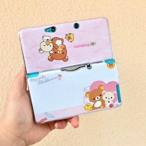 May include: A pink and blue Nintendo 3DS case featuring Rilakkuma characters. The case is open, revealing the screen and buttons. The top half has Rilakkuma and Korilakkuma graphics, while the bottom half has a heart design.