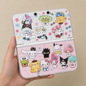 Può includere: Una console di gioco portatile con una custodia protettiva trasparente decorata con vari adesivi dei personaggi Sanrio. L'area dello schermo superiore mostra personaggi come Kuromi e Hello Kitty, mentre quella inferiore ha un motivo a griglia rosa.