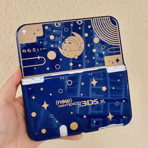 May include: A dark blue New Nintendo 3DS XL handheld gaming console featuring a gold celestial design with stars, planets, and a sleeping character on a crescent moon. The text "ねむたい" and "new NINTENDO 3DS XL" are printed in gold.