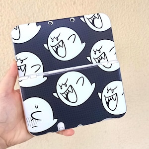 May include: A Nintendo 3DS case with a repeating pattern of white cartoon ghosts on a dark blue background. The ghosts have open mouths and angry expressions. The case is held in a hand.