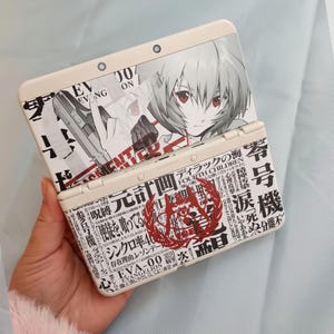 Può includere: Una console di gioco portatile color crema con una grafica anime in bianco e nero. Il design della console include testo giapponese e un emblema rosso. Lo schermo superiore mostra un personaggio con gli occhi rossi.