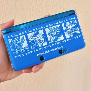 Può includere: Una console di gioco portatile blu con una cover protettiva trasparente. La console presenta illustrazioni grafiche bianche di mostri e il testo "MONSTER HUNTER X". Il design include una serie di triangoli e chevron.
