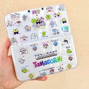 Op de afbeelding: Een witte handheld gameconsole, opengeklapt om een scherm en knoppen te onthullen, is bedekt met kleurrijke cartoonkarakterstickers. De console heeft de tekst "Tamagotchi" en Japanse tekst. Het apparaat wordt in een hand gehouden.