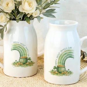 May include: Two white ceramic mugs with handles, each featuring a green rainbow design, a pot of gold, and the text "HAPPY SAINT JAMES'S DAY." One mug holds a bouquet of white roses and greenery. The mugs are on a woven mat.