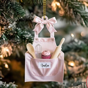 May include: A pink apron-shaped Christmas ornament with a bow at the top, hanging from a gold chain. The apron has a pocket with a wooden spoon, whisk, rolling pin, and a cupcake. The name "Amelia" is printed on the pocket.