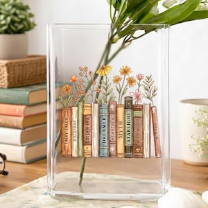 May include: A clear rectangular glass vase with a floral and book design. The design features colorful flowers and a row of books with titles like "Fairy Tales" and "Starlight." The vase holds green stems and white flowers.
