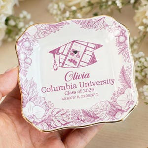 May include: A white ceramic trinket dish with a gold rim, featuring a graduation cap design and the text "Olivia Columbia University Class of 2026". The dish is decorated with a floral border in shades of purple.