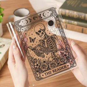 May include: Clear plastic book cover featuring a skeleton reading a book, with a butterfly, roses, and celestial elements. The text "THE READER" is at the bottom. The design is in black and tan.