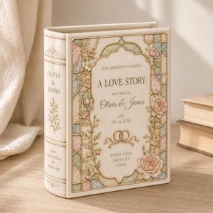 May include: A decorative book-shaped object in cream with pastel floral accents and gold detailing. The cover features the text "OUR GREATEST CHAPTER A LOVE STORY WRITTEN BY OLIVIA & JAMES EST. 09.26.2026" and two interlocking rings. The spine reads "OLIVIA & JAMES" and "THE BEGINNING OF FOREVER."