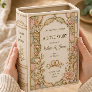 May include: A cream-colored ceramic book-shaped vase featuring a stained-glass style floral border in pastel pink, blue, and green. The front displays the text "OUR GREATEST CHAPTER A LOVE STORY WRITTEN BY OLIVIA & JAMES EST. 09.26.2026" with two gold rings.