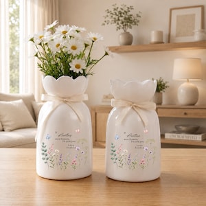 May include: Two white ceramic vases with scalloped rims and cream-colored ribbons tied around the necks. Each vase features a floral print, butterflies, and the text: If Aunties were flowers, I'd pick you. Love Sophia. The larger vase holds white daisies.