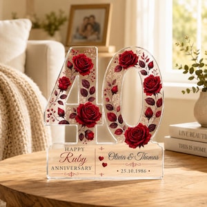 May include: A clear acrylic number 40 sign decorated with red roses and greenery. The text reads: HAPPY Ruby ANNIVERSARY, Olivia & Thomas, and 25.10.1986. The design includes two small red hearts and decorative flourishes on a flat base.