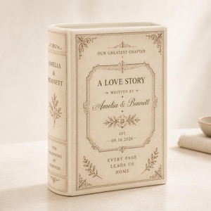 May include: A cream-colored ceramic book-shaped vase featuring brown printed text and floral flourishes. The front reads A LOVE STORY, WRITTEN BY Amelia & Bennett, EST. 08.16.2026, EVERY PAGE LEADS US HOME. The spine displays AMELIA & BENNETT.