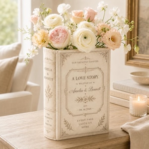 May include: A cream-colored book-shaped vase featuring the text A LOVE STORY, WRITTEN BY AMELIA & BENNETT, EST. 08.16.2026, OUR GREATEST CHAPTER, and EVERY PAGE LEADS US HOME, filled with a bouquet of white, cream, and soft pink ranunculus and sweet pea flowers.
