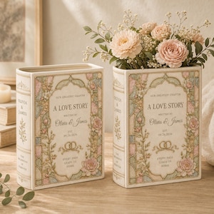 May include: Two rectangular ceramic book-shaped vases featuring a cream background with pastel floral and stained-glass style borders. The text reads: Our Greatest Chapter, A Love Story, Written by Olivia & James, Est. 09.26.2026, and Every Page Leads Us Home.