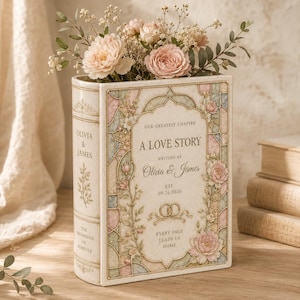 May include: A cream-colored book-shaped vase featuring a stained-glass style floral border in pastel pink, blue, and green. The front displays the text A LOVE STORY, WRITTEN BY OLIVIA & JAMES, EST. 09.26.2026, and two interlocking gold rings.