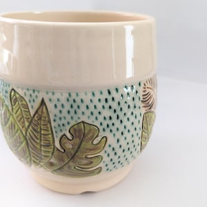 May include: A cream-colored ceramic planter with a tropical leaf design. The planter features a band of green dotted texture and detailed green and brown leaves. The planter is a cylindrical shape.