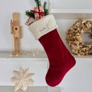 Könnte beinhalten: Ein dunkelroter Weihnachtsstrumpf aus Kord mit einem weißen Sherpa-Umschlag. Auf dem Umschlag befindet sich ein rechteckiges Etikett mit dem Namen "rosalie" in schwarzer Schreibschrift. Der Strumpf ist mit kleinen Geschenken und Tannenzweigen gefüllt.
