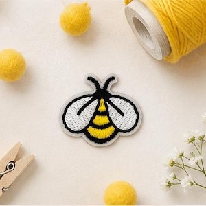 May include: An embroidered bee patch featuring a black and yellow striped abdomen, black antennae, and white wings with black outlines. The patch is shaped like a bee and is displayed as a flat, decorative textile applique for clothing or fabric items.