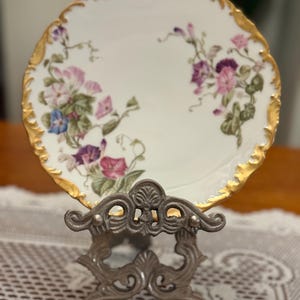 Puede incluir: Un plato de porcelana blanca con un borde festoneado dorado, con un diseño floral en tonos rosa, morado y azul. El plato se exhibe sobre un soporte de metal plateado ornamentado, mostrando una estética vintage.