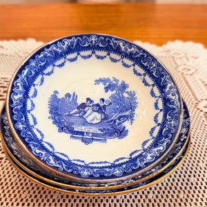 May include: Three blue and white porcelain plates with a gold rim, stacked on a lace doily. Each plate has a blue illustration of a couple in a garden. The plates are a classic design.
