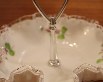 Puede incluir: Una fuente blanca de dos niveles con asa plateada. El plato tiene un borde ondulado con ribete dorado y está decorado con detalles florales morados. Ideal para servir postres o aperitivos.