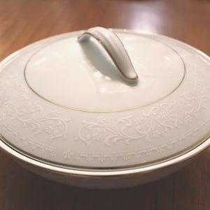 Może przedstawiać: Biała ceramiczna salaterka z pokrywką, z delikatnym kwiatowym wzorem i złotym wykończeniem. Naczynie posiada dwa boczne uchwyty i zakrzywiony uchwyt na pokrywce. Design jest klasyczny i elegancki.