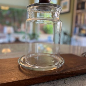 May include: A clear glass cloche with a cylindrical body and a flat circular base. The cloche features a dark brown wooden knob handle on top of the lid. The glass is transparent, allowing a clear view through the container to the surface below.