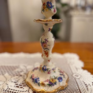 Pode incluir: Um castiçal de porcelana branca vintage com um design floral em azul, amarelo e roxo. As bordas são adornadas com ouro. O castiçal é um elemento decorativo para a casa.