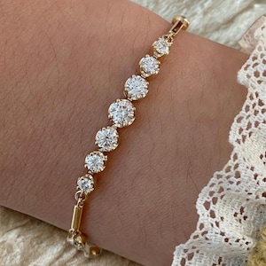 May include: A gold bracelet featuring seven round, clear gemstones. The gemstones are set in individual gold settings and are connected by small gold links. The bracelet is shown on a wrist, with a white lace sleeve visible in the background.