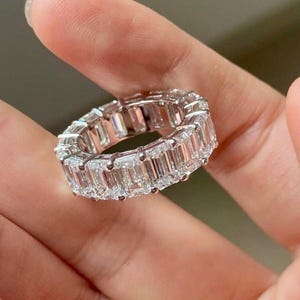 May include: A silver-toned eternity band ring featuring a continuous row of clear, rectangular emerald-cut stones set in a prong setting. The ring encircles the finger, showcasing a uniform arrangement of faceted, sparkling stones around the entire band.